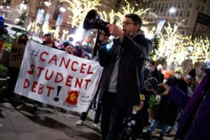 Nighttime rally. Foreground, African American man holding a bullhorn in one hand, gesturing with the other, speaking next to a banner that reads "#Cancel Student Debt." Background, masked rally-goers, and behind them, bare trees strung with lights, and city buildings.