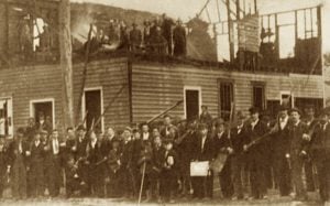 Sepia-toned photo of a crowd of white men posing in front and on top of a half-burned building. The men are wearing suits and white shirts characteristic of the late 19th century. Some are holding rifles.