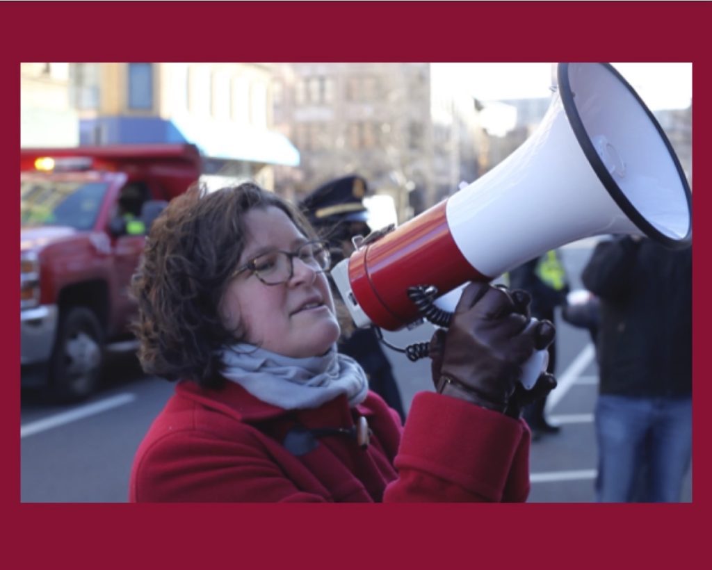 White woman in a red coat and black gloves speaking into a white bullhorn with a red stripe.