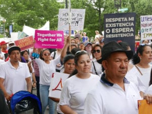 People marching down a tree-lined street. Many wear white T-shirts and carry signs. The sign in the foreground reads “Respeta Mi Existencia o Espera Resistencia,” “Respect My Existence or Expect Resistance.”