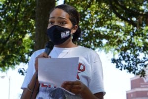 Closeup of a Black woman with warm brown skin speaking into a microphone. Her hair is pulled back; she wears a BLM mask, but the slight drawing-together of her eyebrows and the look in her eyes show deep concern. A bit of a college building shows in the background.