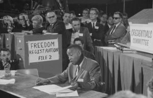 Black-and-white photo. An African American man in a white shirt, suit and tie is seated at a table reading. Several white people stand over him as he does, mostly men, some in sunglasses, expressions ranging from hostile to indifferent. A file cabinet behind the reader to the left has a hand-written sign, “Freedom Registration File 2”; a sign taped to a lectern identifies the credentials committee.