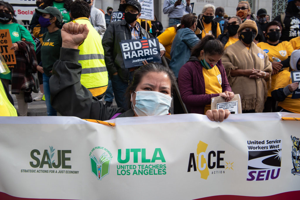 Foreground is a white horizontal banner with organizations’ logos, including UTLA (United Teachers of Los Angeles). In the middle of the frame, a woman holding the banner with one hand, her other fist in the air, defiant expression clear despite her mask. Behind her, a bit of the crowd, mostly Black and Latinx, several wearing orange T-shirts, the color of the inside of a winter squash.