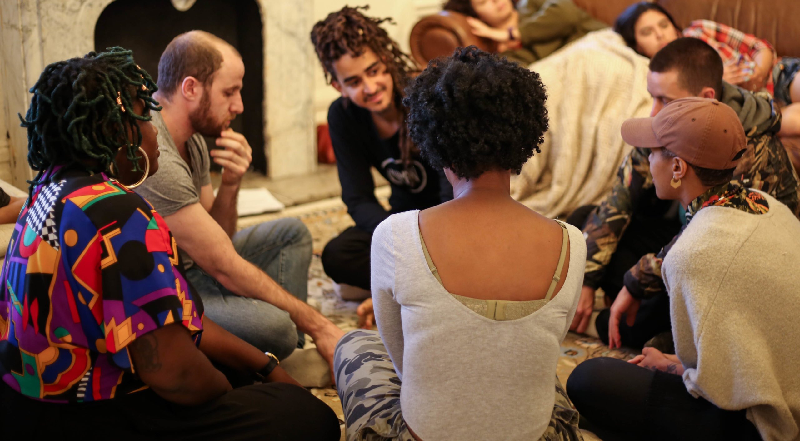 Multi-cultural group of young people sitting in a circle on the floor, leaning in and appearing to listen intently.