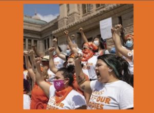 Photo shot from the side. Women wearing white T-shirts and orange bandanas on their heads or around their necks, with their fists in the air. Many are masked but their eyes and body language are fierce.