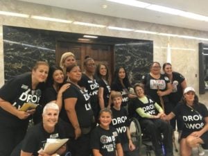Multi-cultural group of women posing in front of a heavy wood door in a marble-walled hallway. They’re wearing black T-Shirts that read “DROP LWOP” in big white letters.