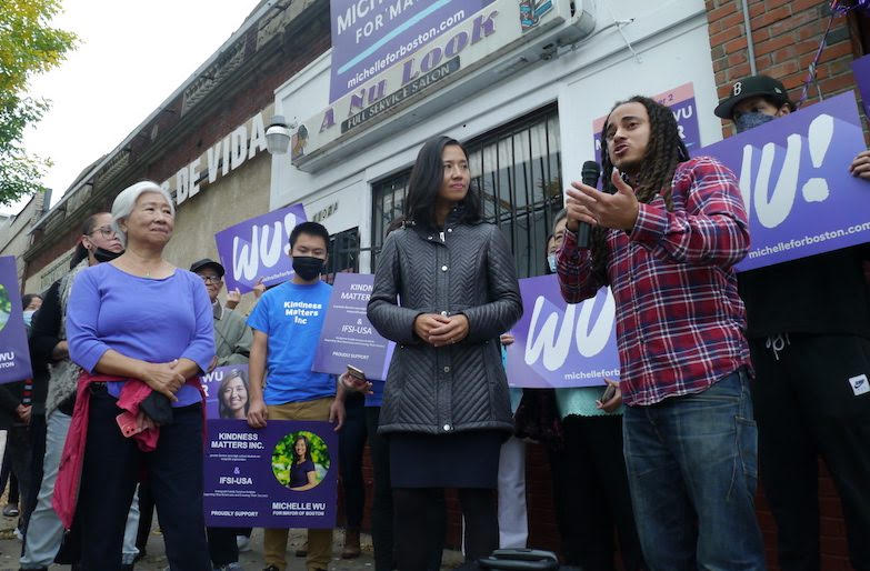Foreground: a brown-skinned man with long black hair holds a microphone and gestures as an Asian American woman looks at him. They’re on the sidewalk in a city small-business district. A small knot of people look on.
