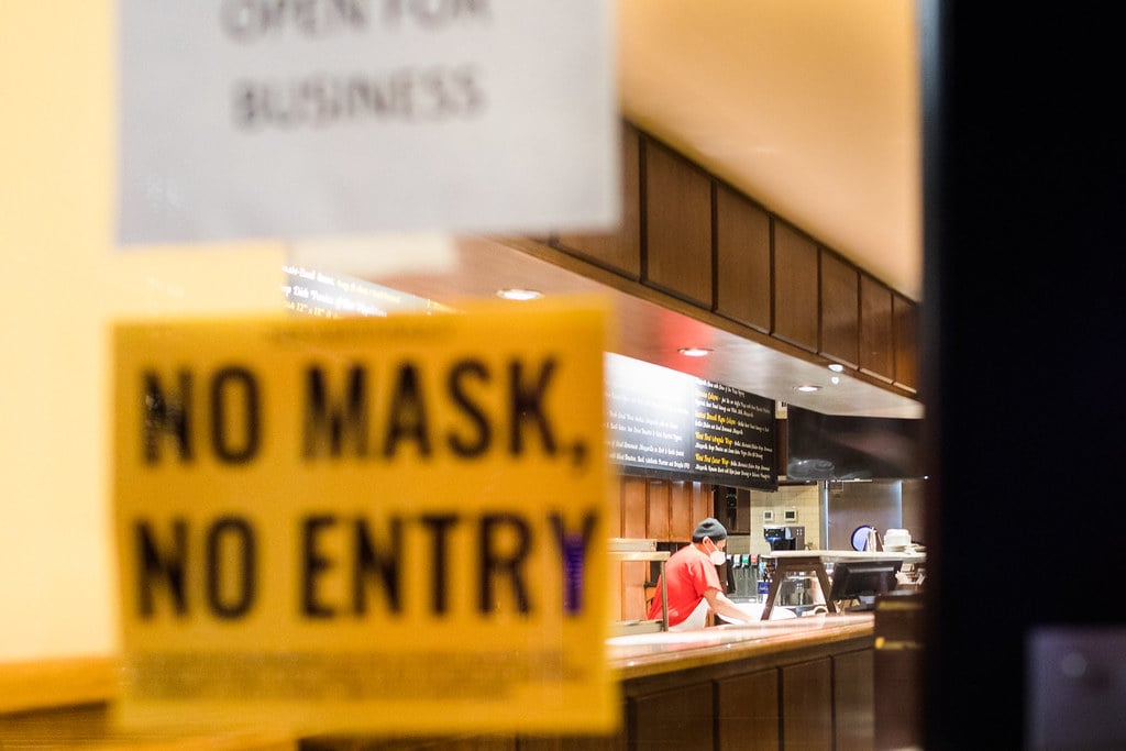 A solitary worker in a mask and apron wipes down a restaurant counter, though the sign on the door says “open for business.”