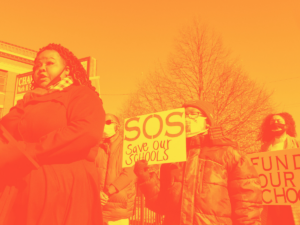 A robust Black woman stands at a makeshift podium in a winter coat; two other women behind her hold signs saying "SOS:Save Our Schools," and "Fund our schools"