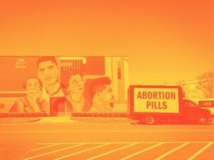 A small black box truck with its side painted white and “ABORTION PILLS” emblazoned on it in huge black letters sits in front of a colorful mural titled “The City of Soul,” featuring four stylized faces.