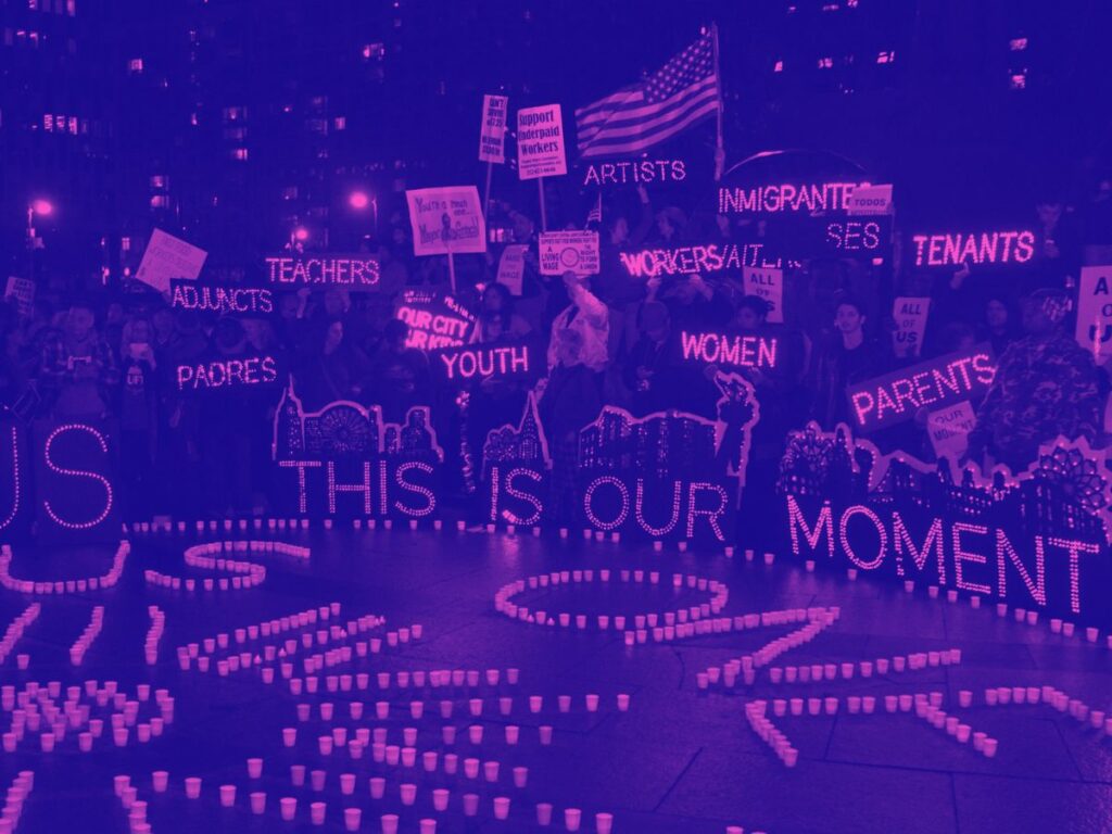 Purple tinted image of protesters holding lit up signs reading encouraging messages with letters made out of lights. Picture is taken at night.