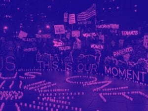 Purple tinted image of protesters holding lit up signs reading encouraging messages with letters made out of lights. Picture is taken at night.