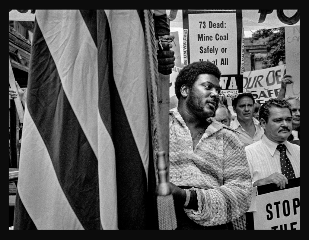 Bob Gumpert photo of Harlan County miners 1974, with a Black man holding a flag in the foreground, UMWA's Harry Patrick in right corner.
