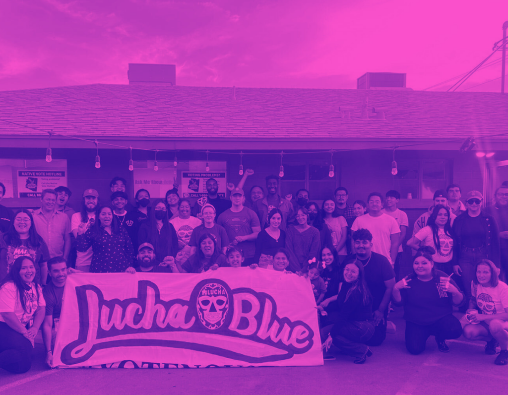Posed photo of a group of people in front of a low building with a banner reading "LUCHA Blue"