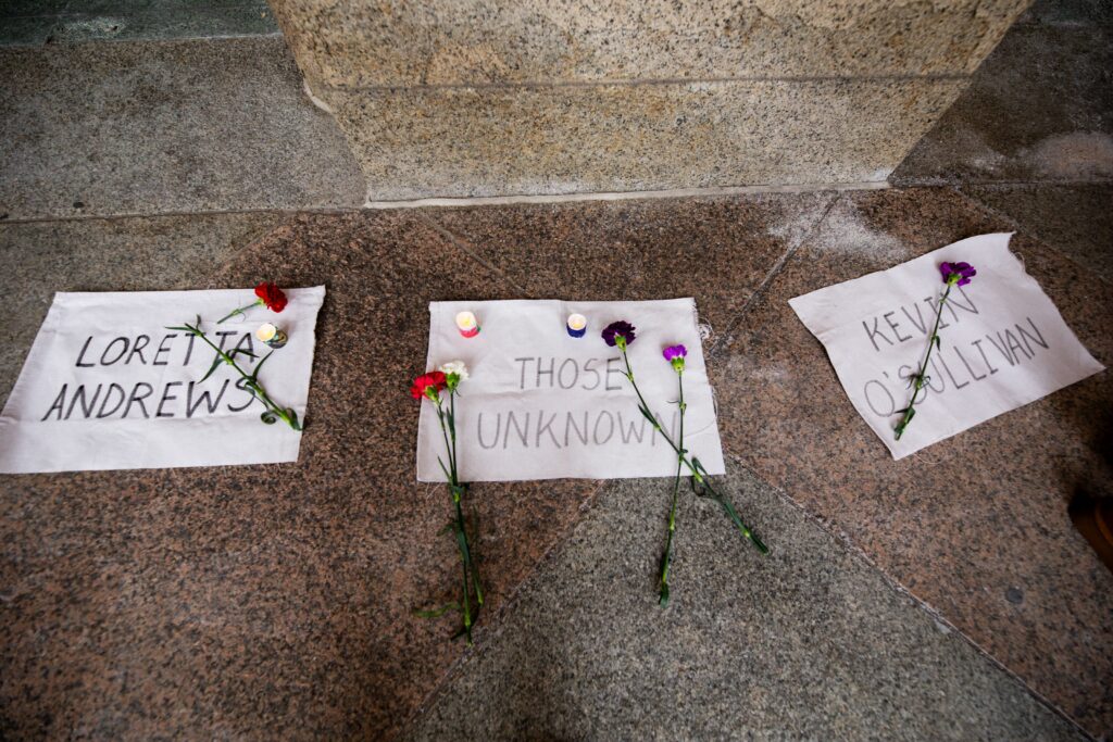 Sheets of paper with names on them, and flowers. placed in a memorial on the sidewalk.